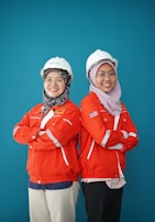 Two individuals are standing back-to-back with arms crossed, wearing red safety jackets and white hard hats. Both are smiling and appear confident. The background is a solid teal color, adding contrast to their bright attire.