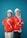 Two individuals are standing back-to-back with arms crossed, wearing red safety jackets and white hard hats. Both are smiling and appear confident. The background is a solid teal color, adding contrast to their bright attire.