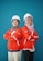 Two individuals are standing back-to-back with arms crossed, wearing red safety jackets and white hard hats. Both are smiling and appear confident. The background is a solid teal color, adding contrast to their bright attire.