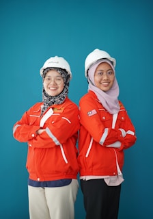 Two individuals are standing back-to-back with arms crossed, wearing red safety jackets and white hard hats. Both are smiling and appear confident. The background is a solid teal color, adding contrast to their bright attire.