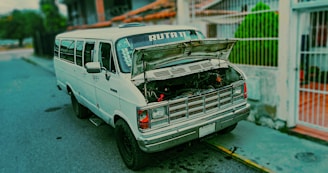 A white van is parked on the side of a street with its hood open, revealing the engine. The van has the word 'RUTA' printed on the front window. It is situated near a residential area with a fence and greenery in the background.