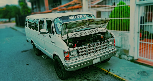 A white van is parked on the side of a street with its hood open, revealing the engine. The van has the word 'RUTA' printed on the front window. It is situated near a residential area with a fence and greenery in the background.