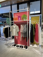 A clothing store display features two racks with various garments. The left rack has patterned shirts and sandals on a shelf below, while the right has dresses in different colors, including pink and black. Decorative elements include orange bottles, a green plant, and a pink backdrop.