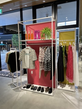 A clothing store display features two racks with various garments. The left rack has patterned shirts and sandals on a shelf below, while the right has dresses in different colors, including pink and black. Decorative elements include orange bottles, a green plant, and a pink backdrop.
