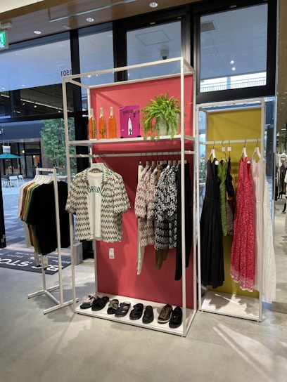 A clothing store display features two racks with various garments. The left rack has patterned shirts and sandals on a shelf below, while the right has dresses in different colors, including pink and black. Decorative elements include orange bottles, a green plant, and a pink backdrop.