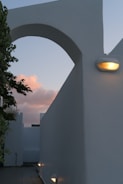 A minimalistic architectural scene features an archway with clean lines and smooth surfaces. The structure is illuminated by a warm light fixture, contrasting against the soft pink and blue hues of the evening sky. Part of a tree with green leaves adds a natural element at the edge of the frame, highlighting the contrast between nature and architecture.