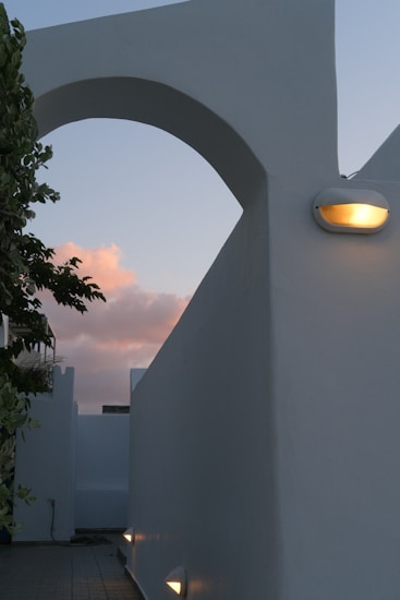 A minimalistic architectural scene features an archway with clean lines and smooth surfaces. The structure is illuminated by a warm light fixture, contrasting against the soft pink and blue hues of the evening sky. Part of a tree with green leaves adds a natural element at the edge of the frame, highlighting the contrast between nature and architecture.