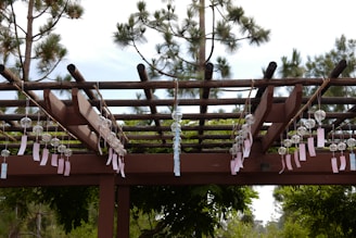 A wooden pergola structure decorated with rows of hanging glass wind chimes, each with a paper tag attached. The background features tall pine trees and clear skies.