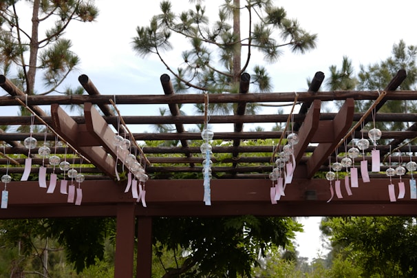 A wooden pergola structure decorated with rows of hanging glass wind chimes, each with a paper tag attached. The background features tall pine trees and clear skies.