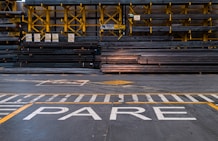 A warehouse or industrial setting with rows of stacked metal beams organized neatly on yellow shelving units. The floor features painted safety lines and a large word 'PARE' in white, likely indicating a stop sign.