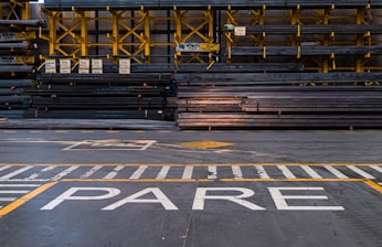 A warehouse or industrial setting with rows of stacked metal beams organized neatly on yellow shelving units. The floor features painted safety lines and a large word 'PARE' in white, likely indicating a stop sign.
