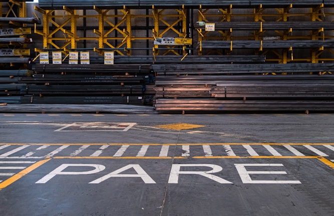 A warehouse or industrial setting with rows of stacked metal beams organized neatly on yellow shelving units. The floor features painted safety lines and a large word 'PARE' in white, likely indicating a stop sign.