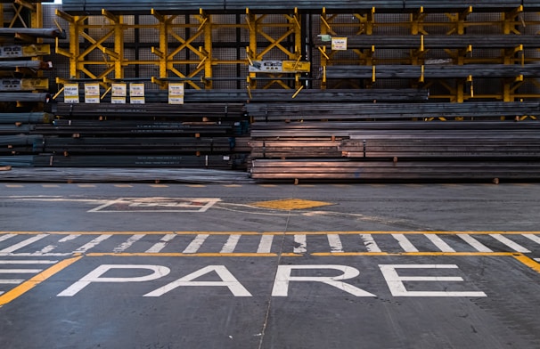 A warehouse or industrial setting with rows of stacked metal beams organized neatly on yellow shelving units. The floor features painted safety lines and a large word 'PARE' in white, likely indicating a stop sign.