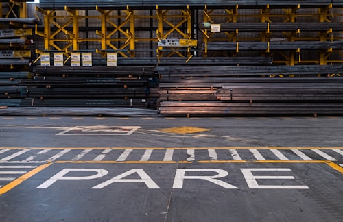 A warehouse or industrial setting with rows of stacked metal beams organized neatly on yellow shelving units. The floor features painted safety lines and a large word 'PARE' in white, likely indicating a stop sign.