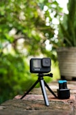 A compact action camera mounted on a small tripod is positioned on a rustic brick surface outdoors. Next to it, two spare batteries are placed in a charger. The background features lush green foliage with a soft, natural light creating a blurred effect.