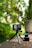 A compact action camera mounted on a small tripod is positioned on a rustic brick surface outdoors. Next to it, two spare batteries are placed in a charger. The background features lush green foliage with a soft, natural light creating a blurred effect.