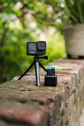 A compact action camera is mounted on a small tripod, placed on a brick wall. Next to the camera is a battery charger containing two batteries. The background is blurred with lush green foliage, suggesting an outdoor setting.