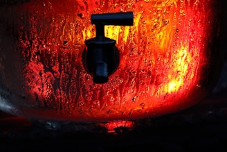 A close-up image of a water faucet or spigot, against a fiery background with red and orange hues. The surface appears wet with condensation, indicating a humid or heated environment. The lighting creates a dramatic and intense effect.