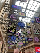 A vibrant indoor space filled with multiple colorful neon signs hanging from a ceiling with a modern architectural design. The signs feature a variety of designs and text in different languages, primarily Chinese. The area is well-lit with a glass roof that allows natural light to filter in.