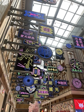A vibrant indoor space filled with multiple colorful neon signs hanging from a ceiling with a modern architectural design. The signs feature a variety of designs and text in different languages, primarily Chinese. The area is well-lit with a glass roof that allows natural light to filter in.