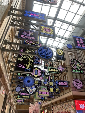 A vibrant indoor space filled with multiple colorful neon signs hanging from a ceiling with a modern architectural design. The signs feature a variety of designs and text in different languages, primarily Chinese. The area is well-lit with a glass roof that allows natural light to filter in.