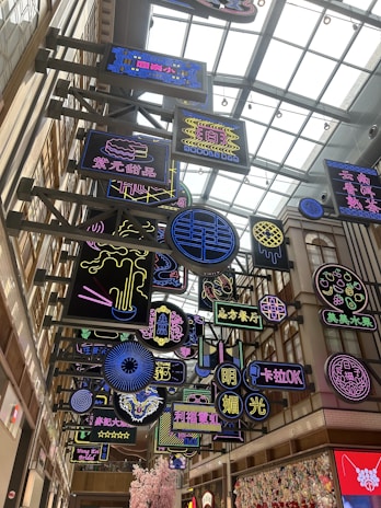 A vibrant indoor space filled with multiple colorful neon signs hanging from a ceiling with a modern architectural design. The signs feature a variety of designs and text in different languages, primarily Chinese. The area is well-lit with a glass roof that allows natural light to filter in.
