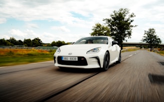 A white sports car is driving down an open road, surrounded by greenery and trees under a partly cloudy sky. The car’s design is sleek and modern, with a visible license plate on the front. The motion of the car suggests speed and freedom as it moves along a scenic route.