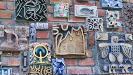 A variety of artistic ceramic tiles are mounted on a brick wall, showcasing intricate designs and patterns. The tiles depict various forms, including geometric shapes, animal figures, and symbolic motifs. The pieces are crafted in different colors and styles, adding a vibrant and eclectic appearance to the wall.