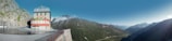 A scenic mountain landscape featuring a building labeled 'Hotel' perched on the edge of a curving road. The background is dominated by lush green mountains and snow-capped peaks under a clear blue sky. The hotel is an isolated structure against the vast expanse of nature, suggesting a remote, peaceful setting.