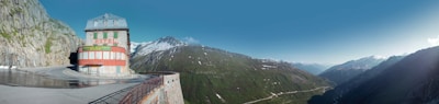 A scenic mountain landscape featuring a building labeled 'Hotel' perched on the edge of a curving road. The background is dominated by lush green mountains and snow-capped peaks under a clear blue sky. The hotel is an isolated structure against the vast expanse of nature, suggesting a remote, peaceful setting.