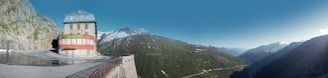 A scenic mountain landscape featuring a building labeled 'Hotel' perched on the edge of a curving road. The background is dominated by lush green mountains and snow-capped peaks under a clear blue sky. The hotel is an isolated structure against the vast expanse of nature, suggesting a remote, peaceful setting.