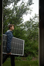 A person is carrying a large solar panel through a lush, green garden area. The environment is rich with greenery, featuring various plants and trees. The lighting suggests an overcast or slightly misty weather, adding a serene atmosphere to the scene.