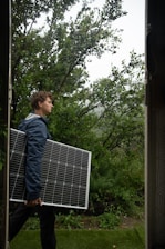 A person is carrying a large solar panel through a lush, green garden area. The environment is rich with greenery, featuring various plants and trees. The lighting suggests an overcast or slightly misty weather, adding a serene atmosphere to the scene.