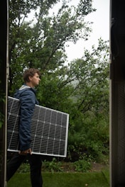 A person is carrying a large solar panel through a lush, green garden area. The environment is rich with greenery, featuring various plants and trees. The lighting suggests an overcast or slightly misty weather, adding a serene atmosphere to the scene.