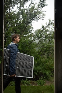 A person is carrying a large solar panel through a lush, green garden area. The environment is rich with greenery, featuring various plants and trees. The lighting suggests an overcast or slightly misty weather, adding a serene atmosphere to the scene.