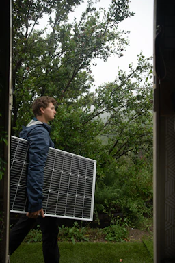 A person is carrying a large solar panel through a lush, green garden area. The environment is rich with greenery, featuring various plants and trees. The lighting suggests an overcast or slightly misty weather, adding a serene atmosphere to the scene.