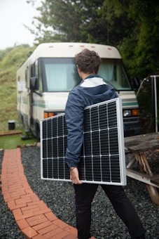A person in a blue jacket carries a large solar panel, walking on a brick pathway surrounded by gravel. In the background, there is an RV parked near greenery.