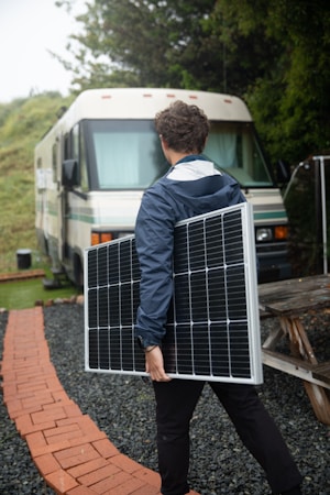 A person in a blue jacket carries a large solar panel, walking on a brick pathway surrounded by gravel. In the background, there is an RV parked near greenery.
