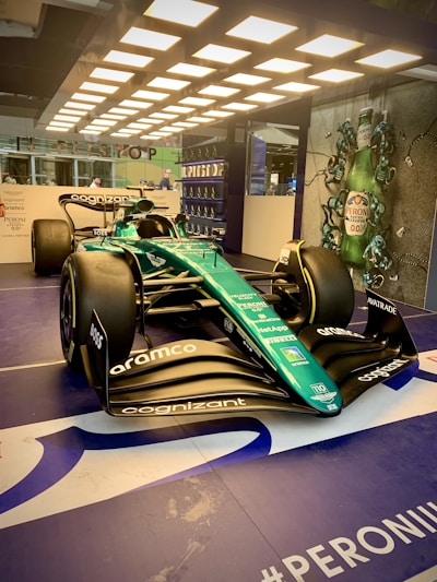 A sleek Formula 1 race car is displayed under bright overhead lights. The car is primarily green with various sponsor logos, including Cognizant and Aramco. In the background, there is a promotional display featuring Peroni beer bottles.