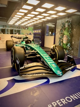 A sleek Formula 1 race car is displayed under bright overhead lights. The car is primarily green with various sponsor logos, including Cognizant and Aramco. In the background, there is a promotional display featuring Peroni beer bottles.