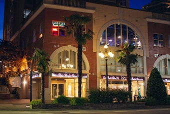A brick building with large arched windows houses a bridal salon. Neon lights illuminate the sign for 'Elks Club' on an upper floor. Several palm trees and a well-lit lamp post are present on the sidewalk. The setting appears to be during the evening, as streetlights are on and the sky is dark.
