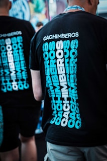 Two people wearing black t-shirts with the word 'Cachimberos' written multiple times in a bold, blue font, standing in a casual setting. The background is out of focus, suggesting an indoor or festival environment.