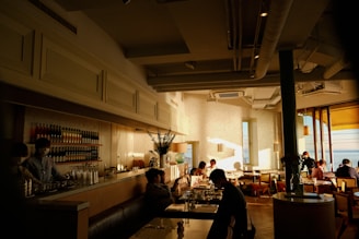 A cozy cafe with a warm, inviting atmosphere. Sunlight streams through large windows, creating a pattern of light and shadow on the white brick walls. Several people are seated at tables enjoying conversation and drinks. There is a well-stocked bar in the background, with bottles neatly arranged on shelves. The decor is modern, with a mix of natural and artificial light.