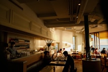 A cozy cafe with a warm, inviting atmosphere. Sunlight streams through large windows, creating a pattern of light and shadow on the white brick walls. Several people are seated at tables enjoying conversation and drinks. There is a well-stocked bar in the background, with bottles neatly arranged on shelves. The decor is modern, with a mix of natural and artificial light.