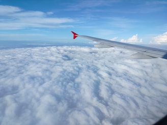 An airplane wing is visible above a vast expanse of white clouds, with a clear blue sky meeting the horizon. The wing is sleek and metallic, with a red tip standing out against the white and blue backdrop.