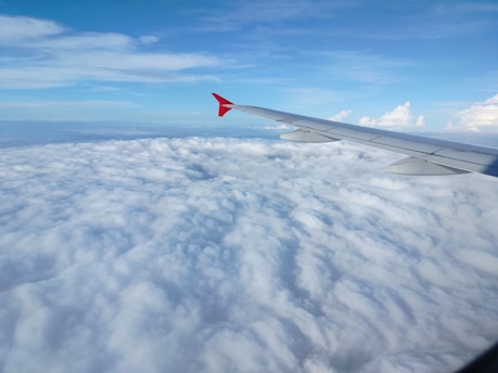 An airplane wing is visible above a vast expanse of white clouds, with a clear blue sky meeting the horizon. The wing is sleek and metallic, with a red tip standing out against the white and blue backdrop.