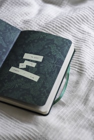 An open notebook with a dark green cover featuring a pattern of pine leaves. Inside, there are small, rectangular labels with text on them. A green ribbon bookmark is visible, and the book is resting on a textured, white fabric surface.