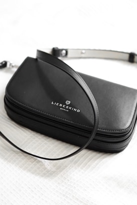 A sleek, black leather handbag with a silver strap featuring a simple and elegant design. The brand name 'Liebeskind Berlin' is imprinted on the front of the bag, which rests on a textured white surface.