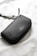A sleek, black leather wallet or clutch with a simple, elegant design and a brand logo embossed on its surface is placed on a textured white fabric background. A detachable strap is partially visible, suggesting versatility and functionality.