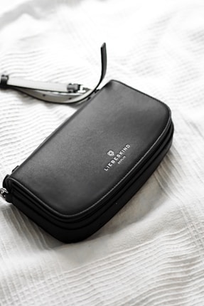 A sleek, black leather wallet or clutch with a simple, elegant design and a brand logo embossed on its surface is placed on a textured white fabric background. A detachable strap is partially visible, suggesting versatility and functionality.